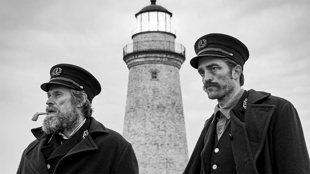 Willem Dafoe e Robert Pattinson em personagens com um farol ao fundo em The Lighthouse. 