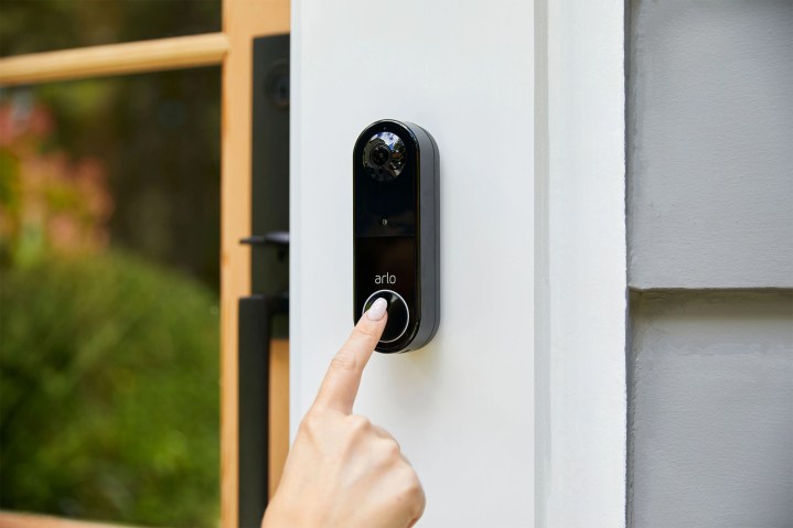 A finger touching the Arlo Video Doorbell, mounted on a house wall.