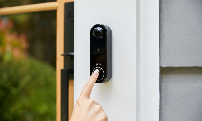 A finger touching the Arlo Video Doorbell, mounted on a house wall.
