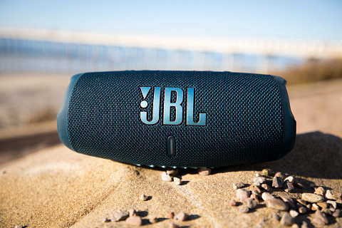 A JBL Charge 5 Bluetooth speaker sits on a beach.