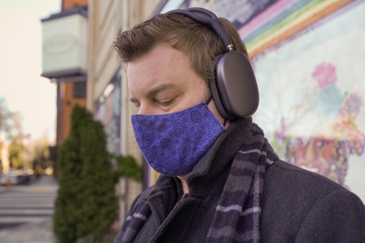 A man wearing Apple AirPods Max as well as a mask, while sitting against a wall.