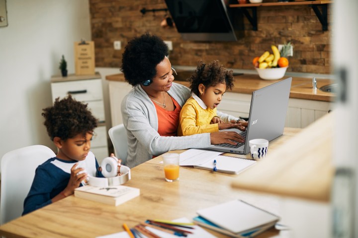 A Black family works on various projects at a dining table. A mother with daughter on her lap works at an Acer computer while a boy adjusts headphones. 