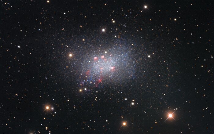 Sextans B is an irregular dwarf galaxy, meaning that it is irregularly shaped and smaller than our galaxy, the Milky Way. It lies around 4.5 million light-years from Earth and is located in the constellation Sextans in the southern sky. Captured with the Nicholas U. Mayall 4-meter Telescope, this image of Sextans B features red-colored star-forming regions near the galaxy’s center. Surrounding the galaxy are several bright stars that are located much closer to us in our galaxy, identified by the crisscross patterns created by light interacting with the structure of the telescope, as well as numerous fuzzy-looking background galaxies that appear small because they are much farther away than Sextans B.