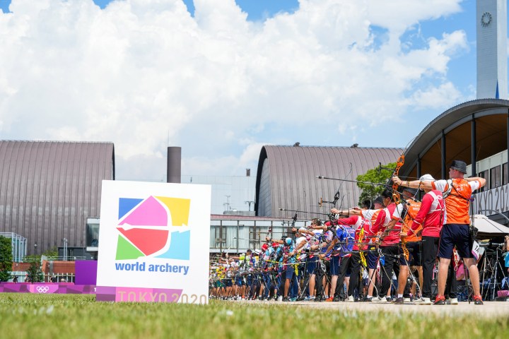 A group of archers with bows drawn aim over a sign for World Archery Tokyo 2020.