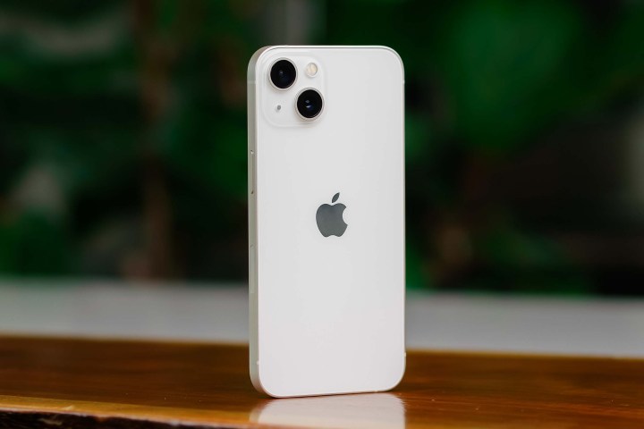 Белый iPhone 13 стоит вертикально на столе.