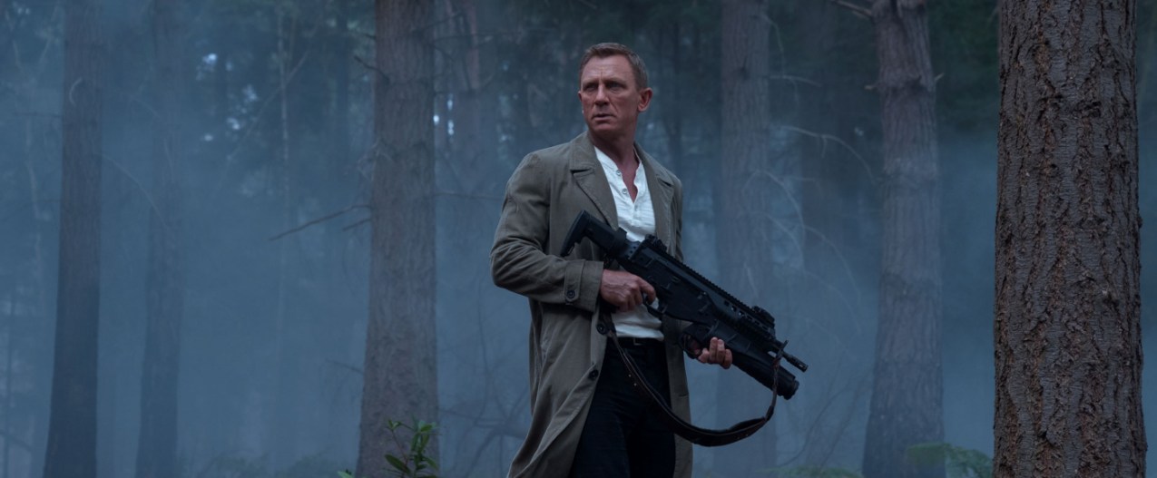 Daniel Craig stands in a forest with a rifle in a scene from No Time To Die.
