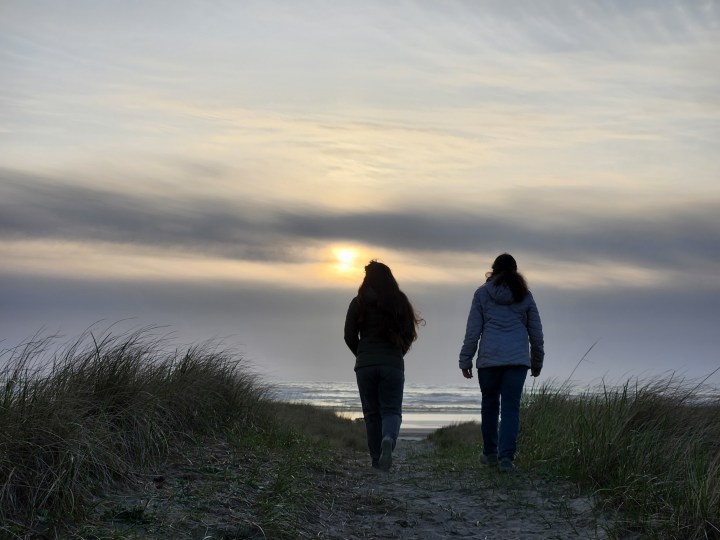 Two people walking over the top of a dune with the ocean and sunset in the background.