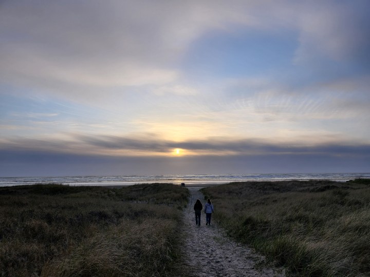Sunset over dunes with grassy fields and two people walking on a path towards the ocean.