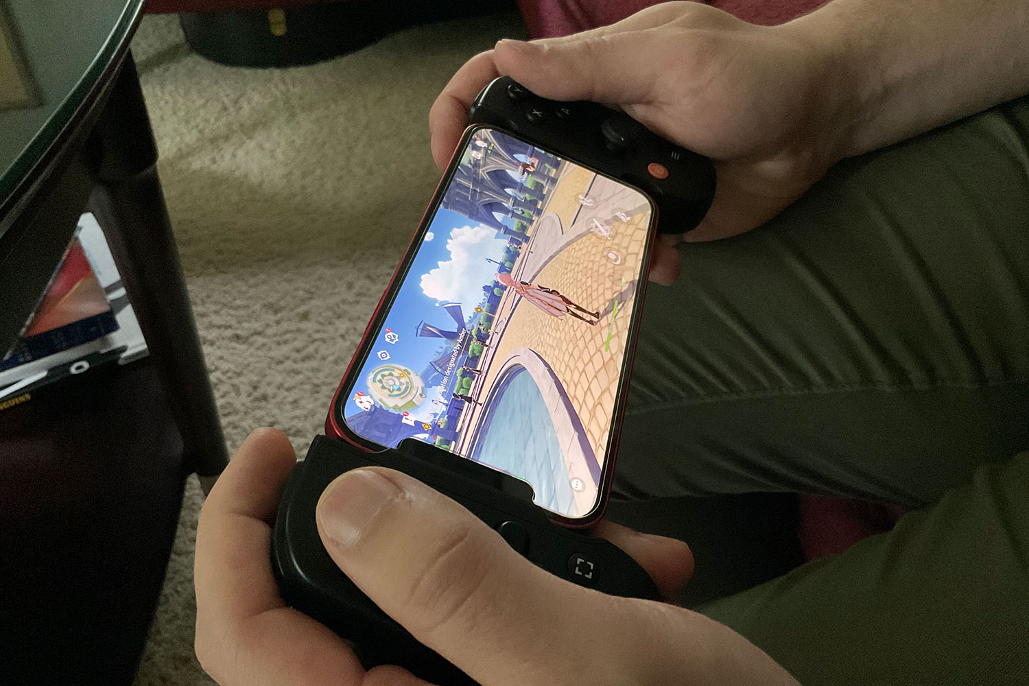 A Backbone controller held in a person's hands with an iPhone 12 inside it. On the screen of the phone, Genshin Impact is being played.