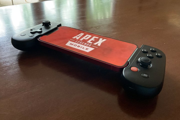 A Backbone controller with an iPhone 12 inside it. The devices are resting on a table and the Apex Legends Mobile logo is displayed on the phone's screen.