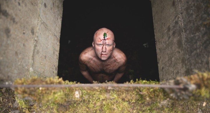 Rory Kinnear looks up, naked, from a pit, with scratches on his body and a leaf on his head.
