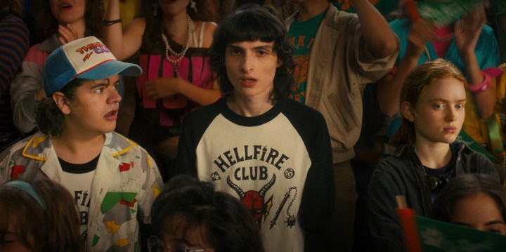 Gaten Matarazzo, Finn Wolfhard et Sadie Sink se tiennent dans la foule lors d'un rassemblement au lycée dans une scène de Stranger Things 4.