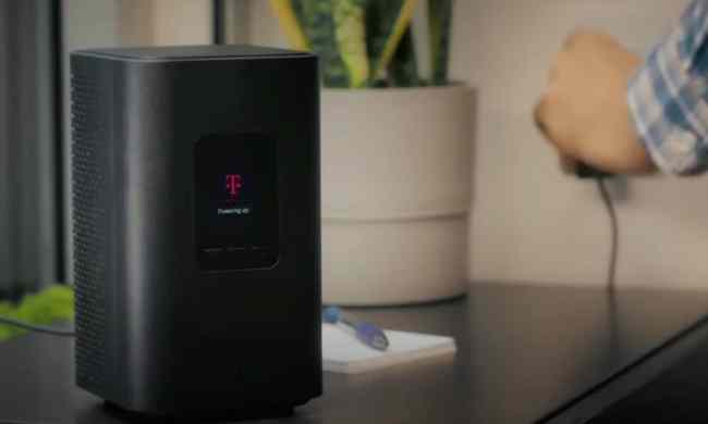 T-Mobile 5G home internet router on tabletop with a person's hand plugging in the power adapter in the background.