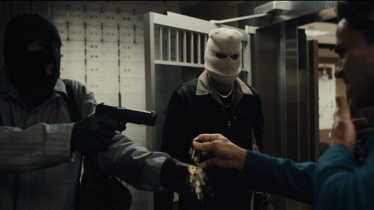Três ladrões de banco seguram um trabalhador sob a mira de uma arma em uma cena de Blowback.