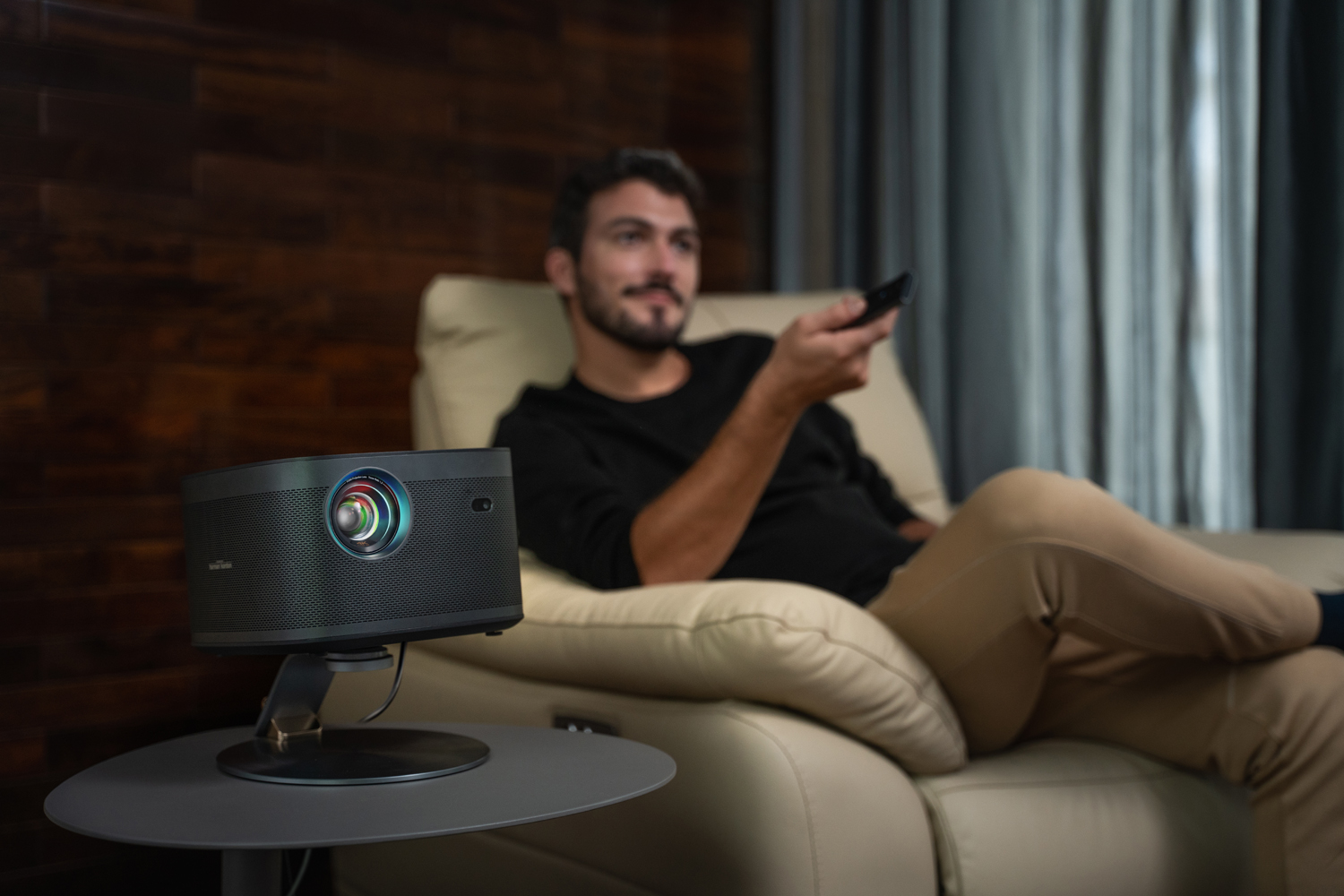 Un hombre en una silla con mando a distancia y un proyector sobre una mesa. 