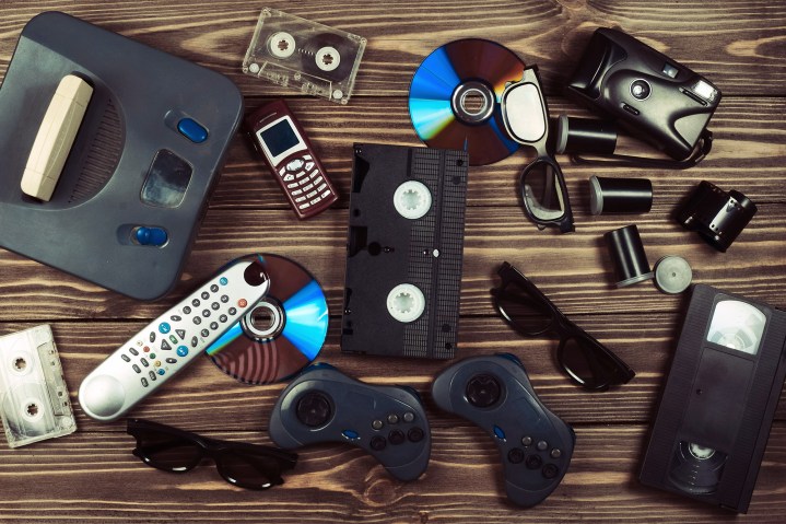 Vintage media and information technology. Entertainment 90s. Game console, gamepads, disks, audio cassettes, video cassettes, phone, film camera on a wooden table.