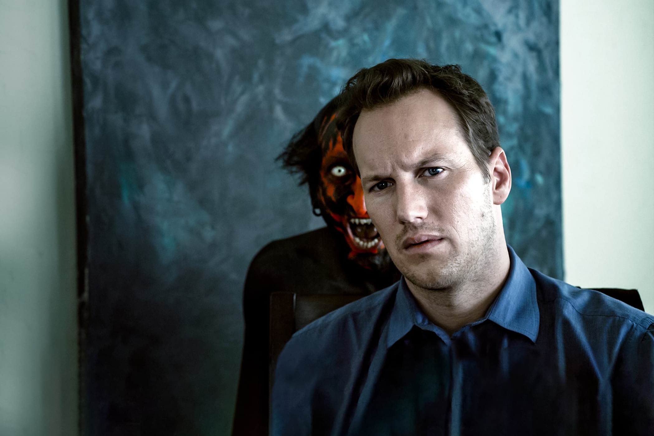 O personagem principal de Insidious está sentado em uma sala olhando para frente enquanto um assustador demônio de rosto vermelho aparece atrás dele rosnando.