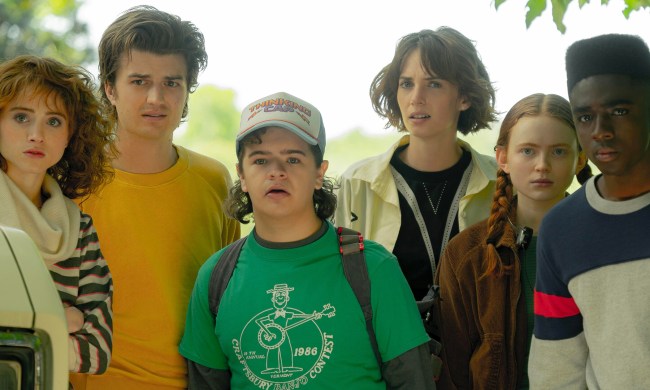 The cast of Stranger Things season 4 is standing in front of a field looking surprised.