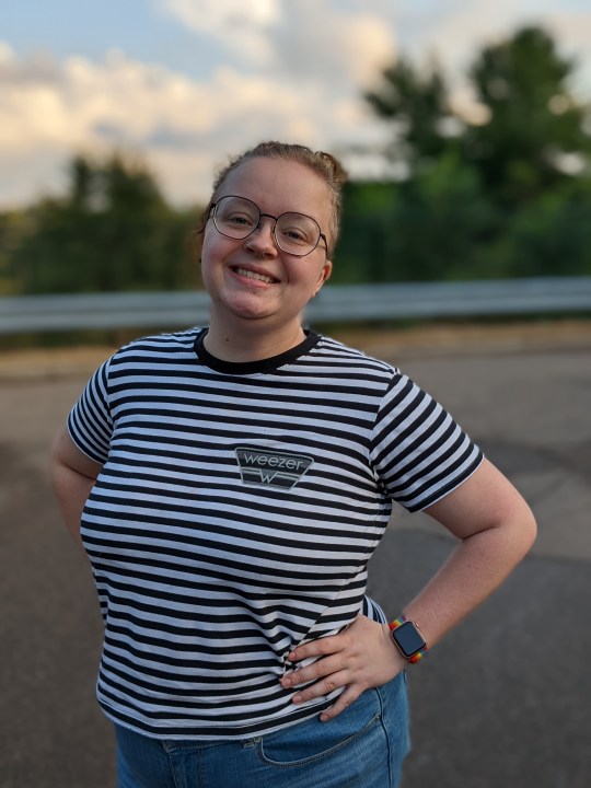 A person with blond hand, tied up in a bun. They're wearing a back and white striped shirt, hands on hips, and standing outside with a smile on their face.