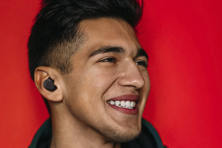 A smiling young man with dark hair wears the JBL Vibe 100 TWA earbuds in his ears.