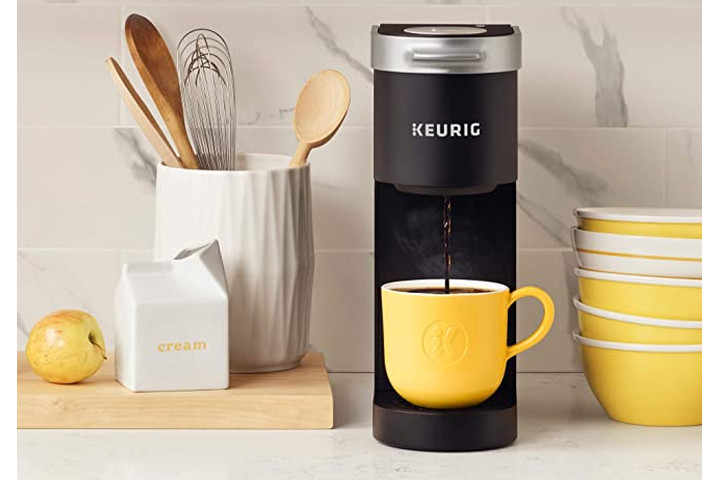 The Keurig K-Mini Coffee Maker brews coffee in a yellow mug on a kitchen counter, next to a tub of kitchen utensils.