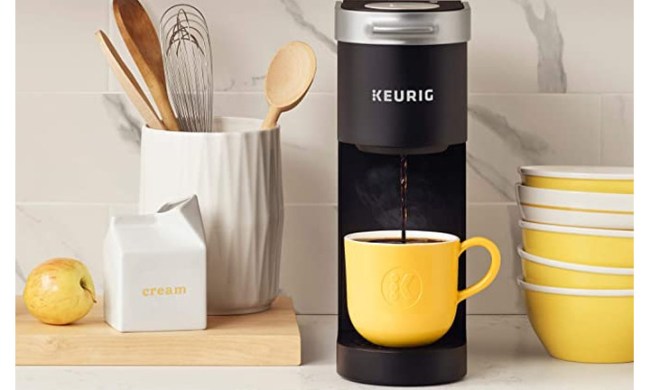 The Keurig K-Mini Coffee Maker brews coffee in a yellow mug on a kitchen counter, next to a tub of kitchen utensils.