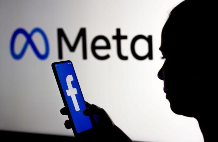 A silhouetted person holds a smartphone displaying the Facebook logo. They are standing in front of a sign showing the Meta logo.