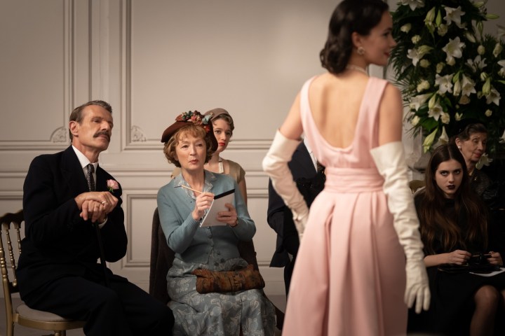 As pessoas olham para uma modelo posando com um vestido rosa em Mrs. Harris Goes to Paris.