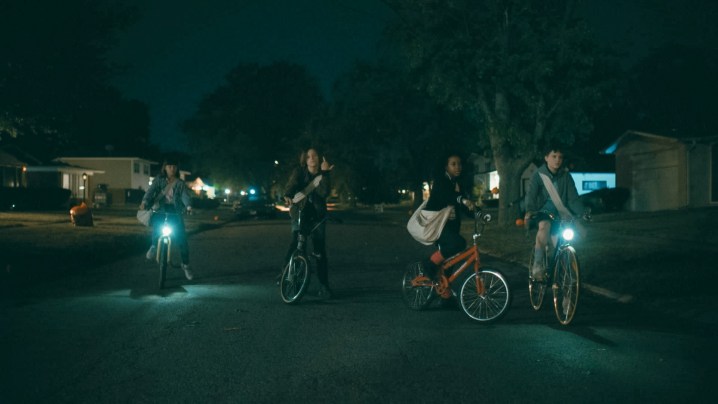 As quatro estrelas de Paper Girls sentam em suas bicicletas em uma rua escura.