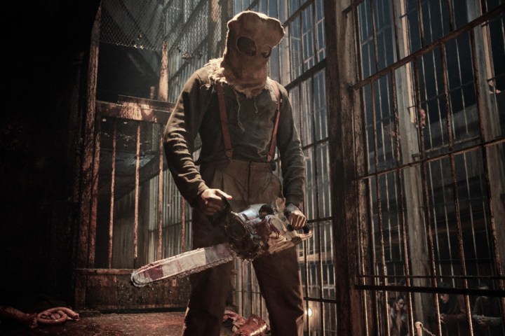 A hooded figure holding a bloody chainsaw stands amid prison cells in a scene from the Resident Evil series.