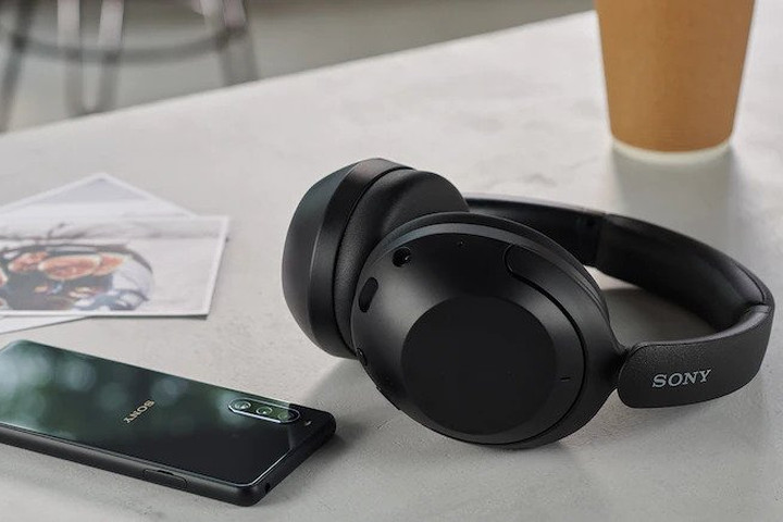 Sony's WH-XB910N wireless noise-cancelling over the ear headphones lie on a table next to a smartphone.