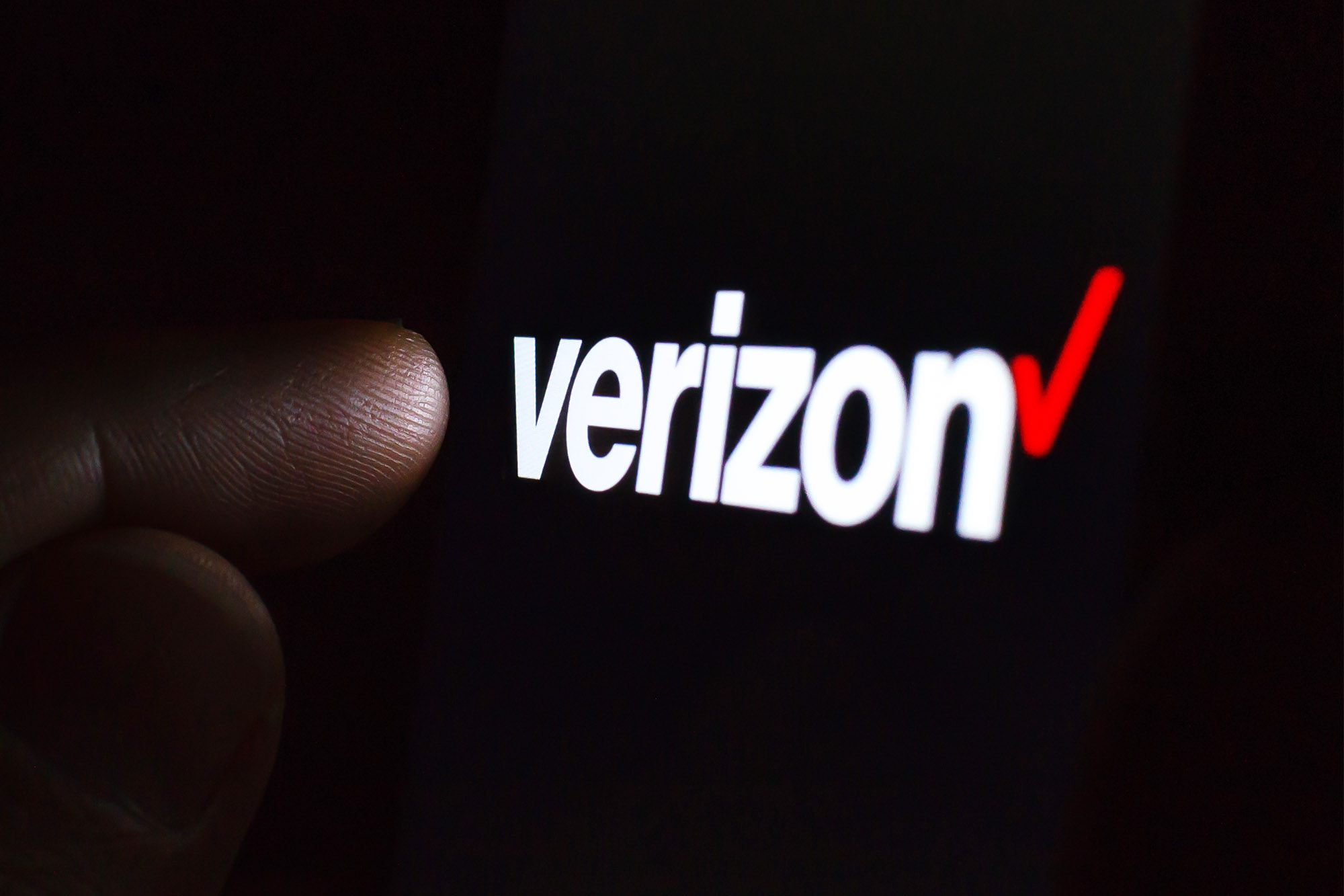 لوگوی Verizon روی صفحه گوشی هوشمند در اتاقی تاریک و انگشتی که آن را لمس می کند.