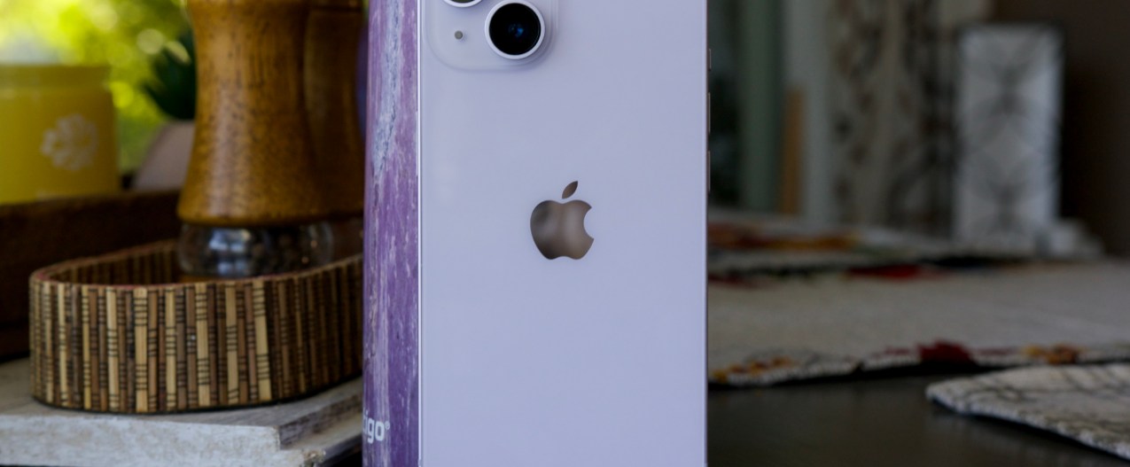 An iPhone 14 standing up on a table. Behind it is a purple water bottle.