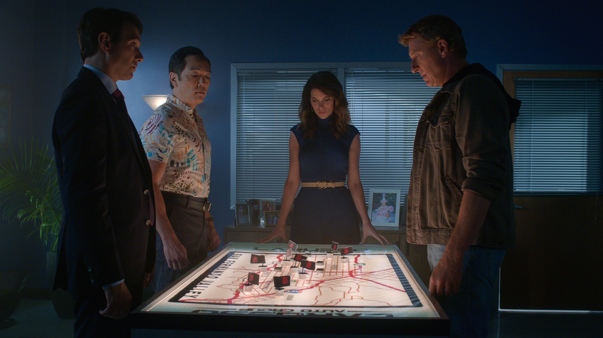 Ralph Macchio, Yuji Okumoto, Courtney Henggeler und William Zabka stehen in einer Szene aus Staffel 5 von Cobra Kai um einen Tisch herum.