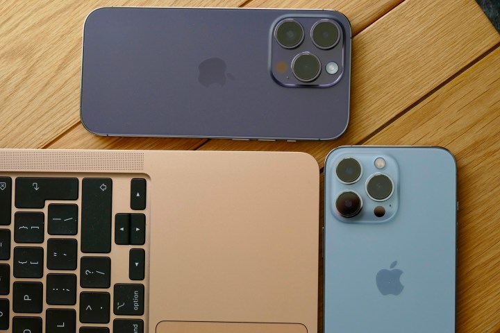 The iPhone 14 Pro and iPhone 13 Pro face down on a table with a MacBook Air.