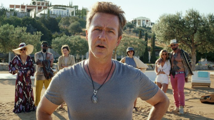 Edward Norton stands on a beach with people behind him in Knives Out 2.