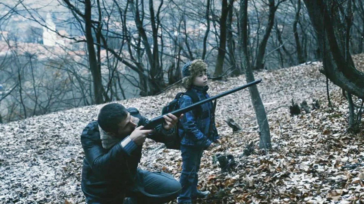R.M.N में एक आदमी अपने बगल में एक लड़के के साथ एक बंदूक का लक्ष्य रखता है।
