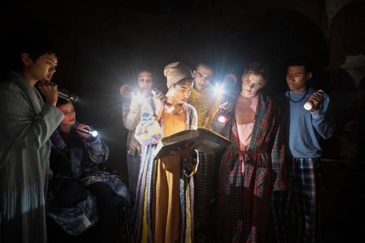 The cast of The Midnight Club reads a book with a flashlight.
