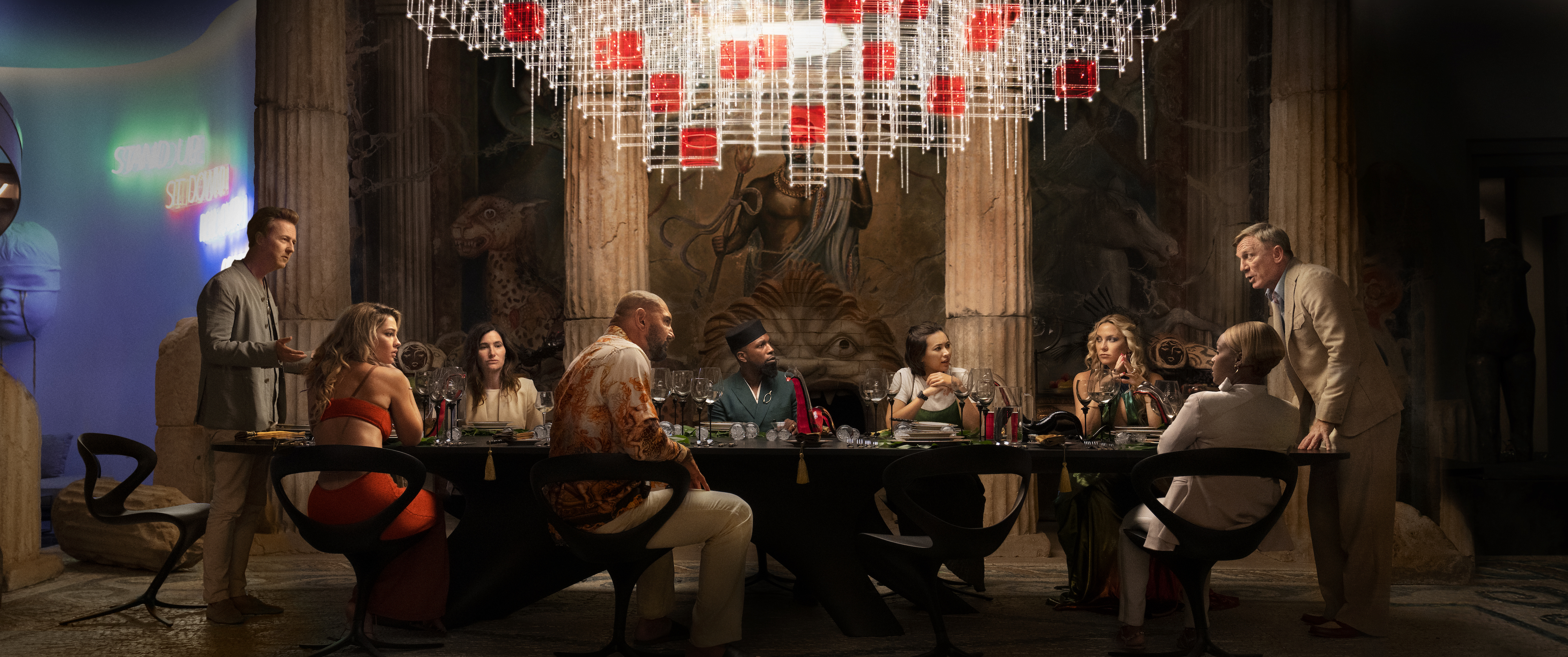 O elenco de Glass Onion se reúne em torno de uma mesa em uma cena do filme da Netflix.
