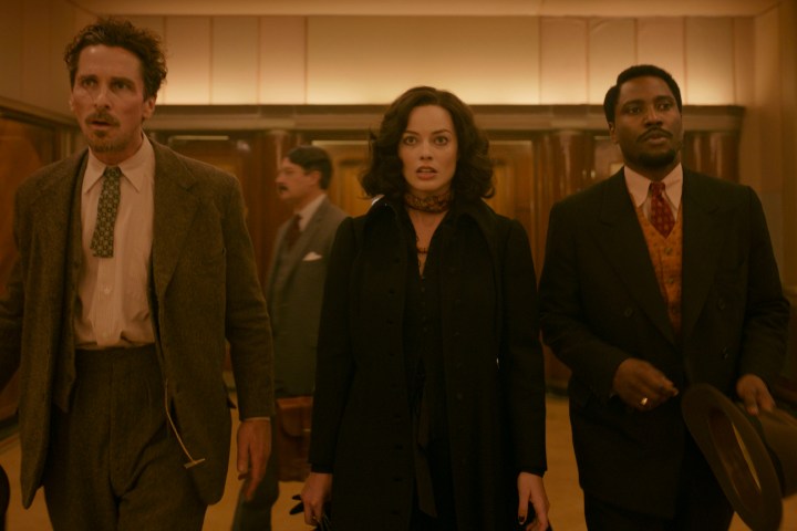 Christian Bale, Margot Robbie und John David Washington gehen gemeinsam durch eine Halle in Amsterdam.