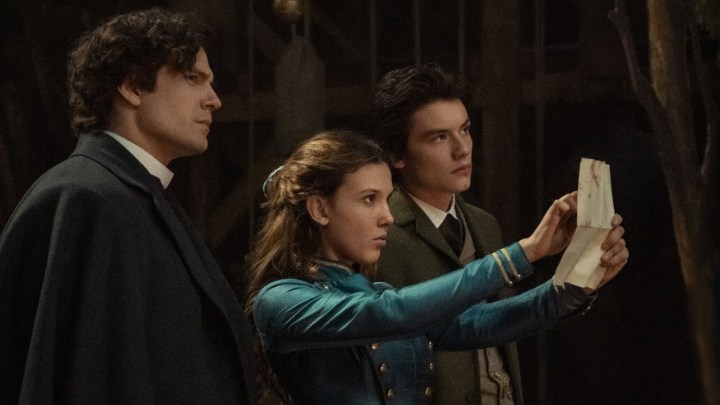 Millie Bobby Brown, Henry Cavill, and Louis Partridge as Sherlock, Enola, and Tewkesbury staring at a piece of paper in a scene from Enola Holmes 2.