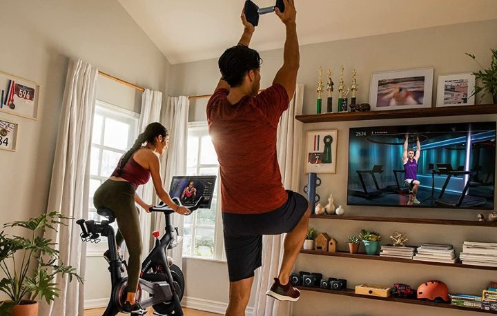 Woman riding original Peloton Bike in a family room with a man working out with weights with interactive class.