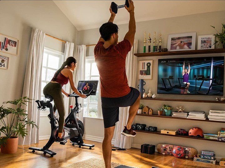Woman riding original Peloton Bike in a family room with a man working out with weights with interactive class.