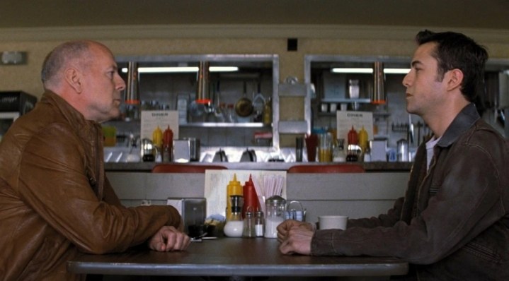 A man sits at a table with a younger man in Looper.