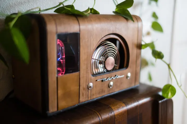 Una PC dentro de una caja de radio Motorola de madera vintage con un ventilador de acero inoxidable en la parte delantera, rodeada de hojas verdes.