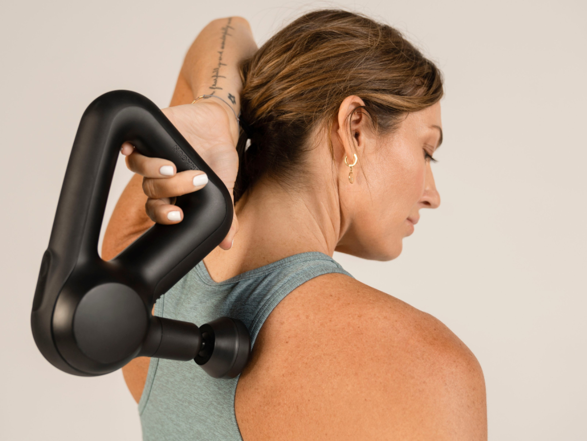 Женщина использует портативное устройство для перкуссионного массажа Theragun Prime на верхней части спины.