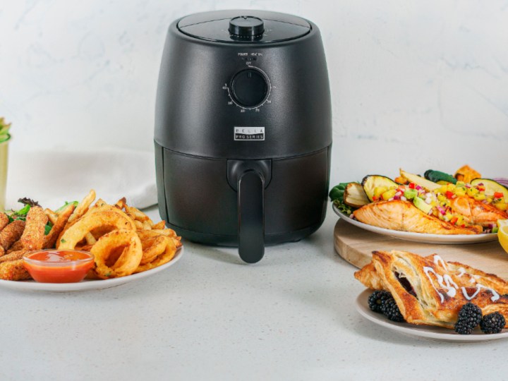 Bella Pro Series - 2-quart analog air fryer on a counter with several dishes of cooked food.