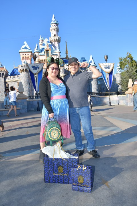 Christine and her husband pose in front of the Disneyland castle with shopping bags from Club 33.