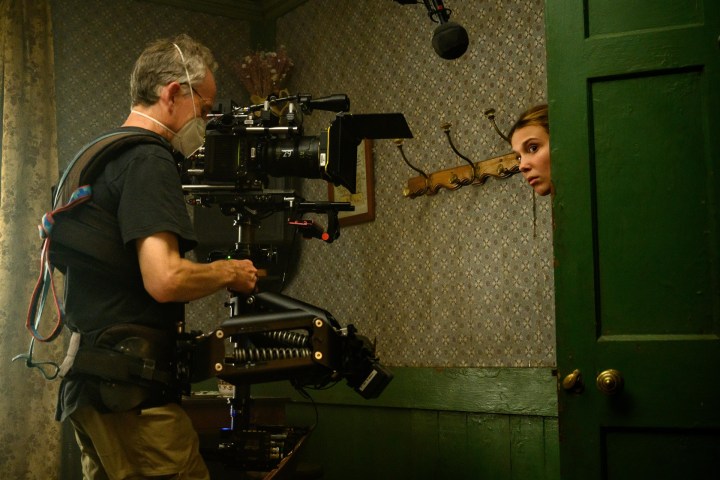Millie Bobby Brown peeks out the door while being photographed in a behind-the-scenes photo of Enola Holmes 2.