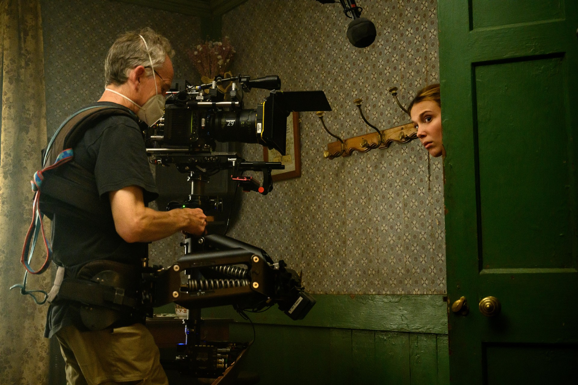Millie Bobby Brown espia por uma porta enquanto é filmada em uma foto dos bastidores de Enola Holmes 2.